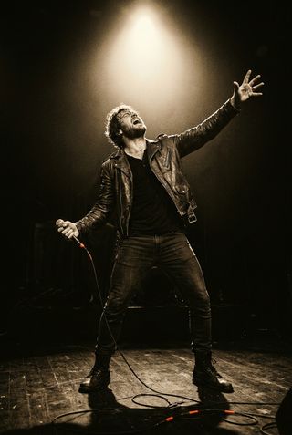 Singer performing on stage under a spotlight, arm outstretched to the crowd
